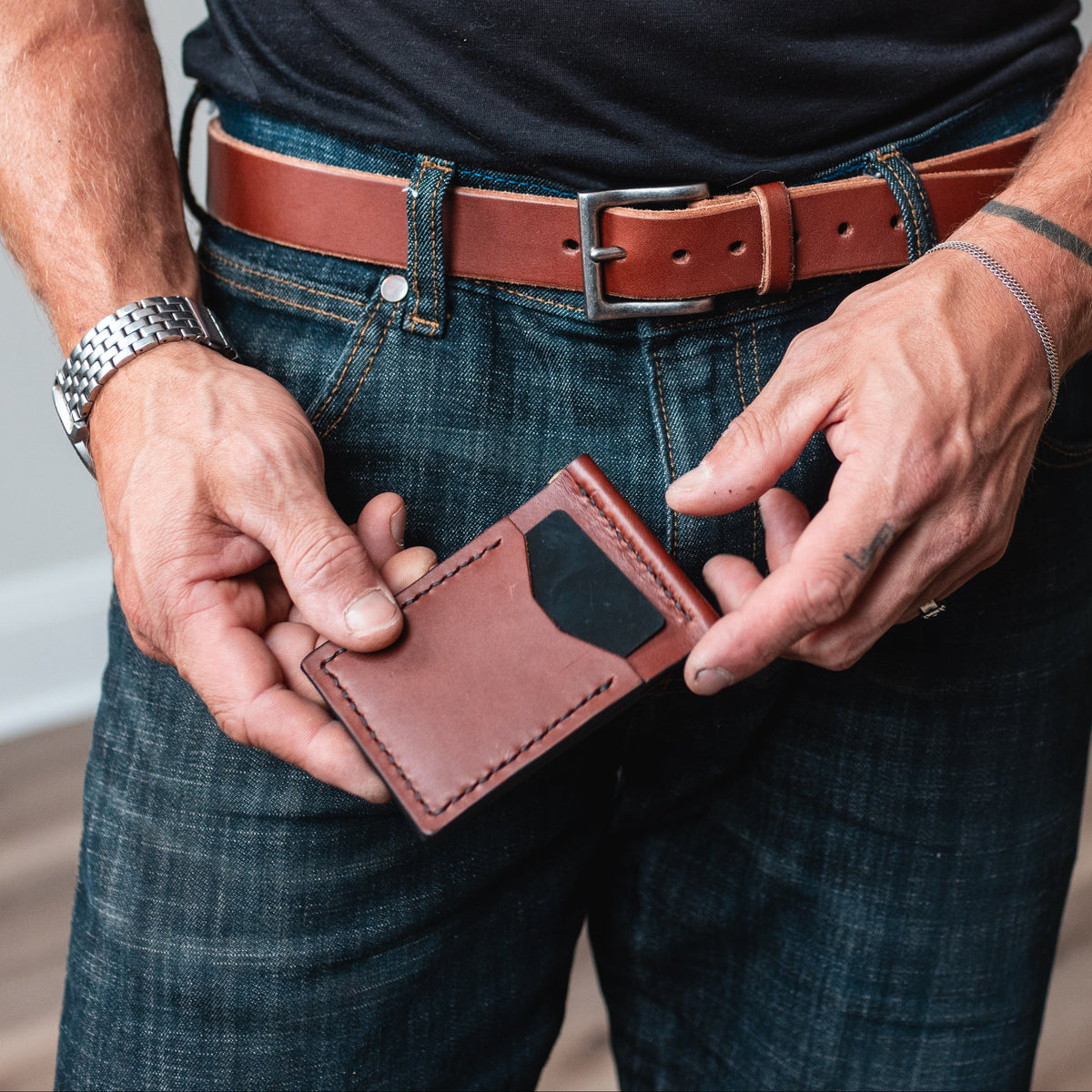 A man wearing a black t-shirt and blue jeans holds a slim cognac brown leather money clip card holder wallet in front of his waist. He is wearing a matching leather belt 