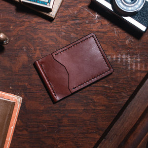 A cognac brown leather card holder wallet with visible stitching, placed on a dark wooden desk surrounded by vintage props including a camera, a brass object, and a book. Handmade by Jesse Bateson in Peterborough Ontario Canada.