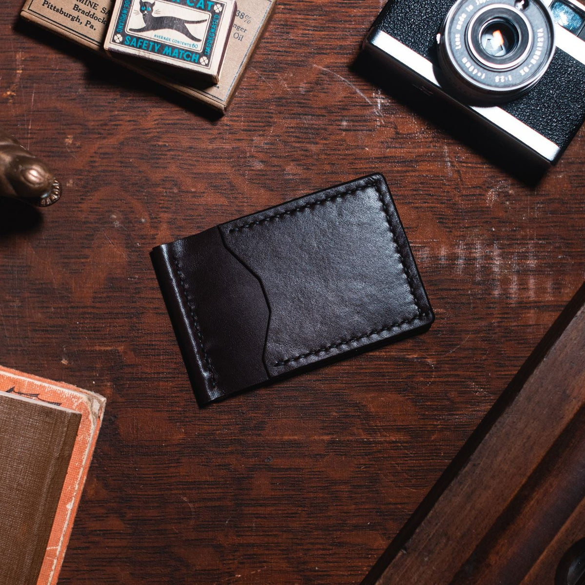 A dark brown solid leather card holder wallet with contrast stitching, displayed on a dark wooden desk surrounded by vintage props including a camera, a small metal alligator figurine, and a textured book.