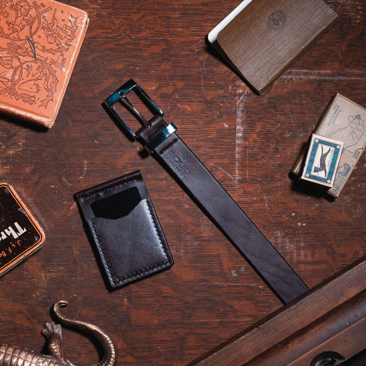A dark brown formal solid leather belt with a stainless steel buckle and keeper  matching dark brown solid leather card holder wallet, arranged on a rustic dark wooden table alongside other vintage leather goods.