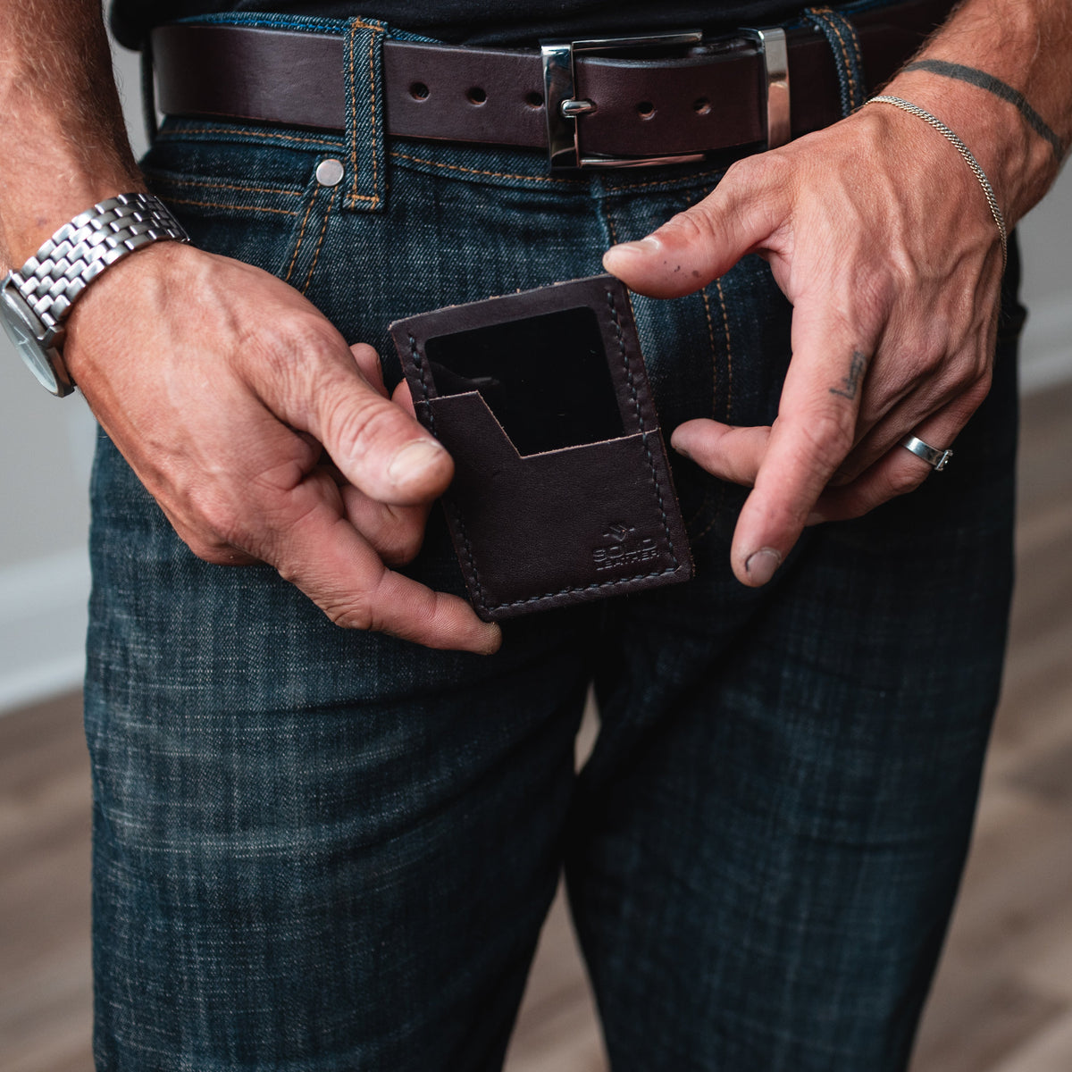 a pair of mans hands holds a dark brown leather cardholder wallet with a matching full-grain brown leather belt on blue jeans. 