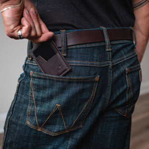 A close-up lifestyle shot of a person wearing dark blue denim jeans with a dark brown full-grain leather belt and a matching brown leather card holder wallet. 