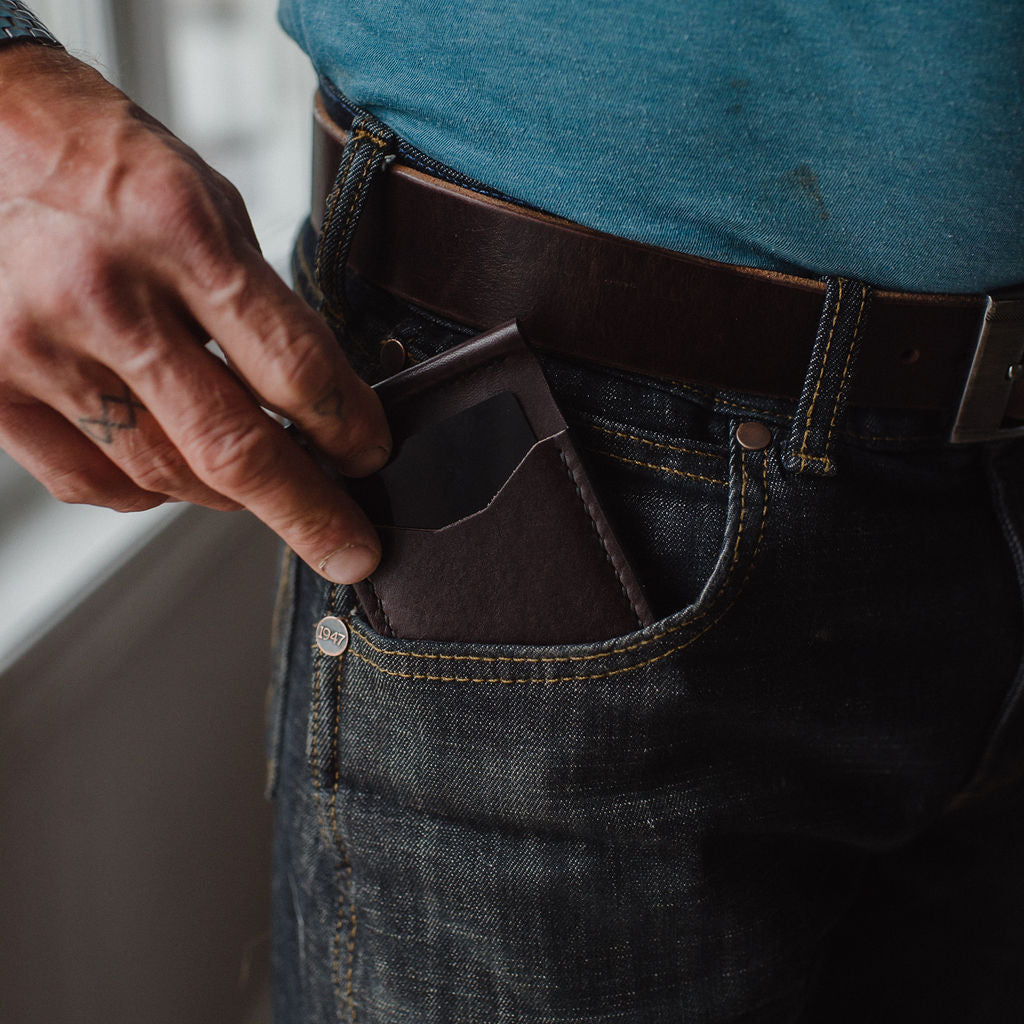 A close-up lifestyle shot of a man's hand sliding a dark brown solid leather card holder wallet into the pocket of his dark blue jeans, wearing a blue t-shirt and a brown leather belt Handmade in Peterborough Ontario Canada by Jesse Bateson. 