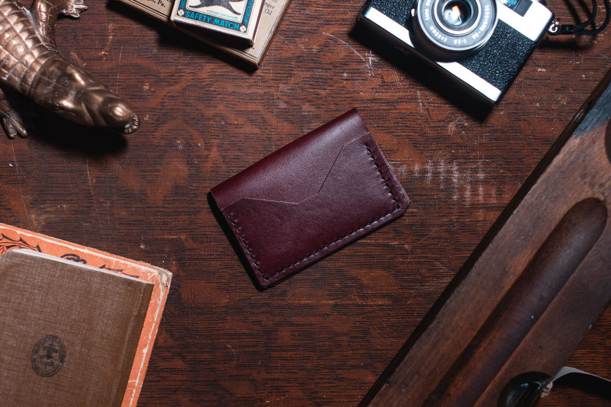 A dark burgundy leather card holder wallet with a stitched envelope-style flap, placed on a dark wooden tabletop surrounded by vintage props including a camera, notebook, and metal object.
