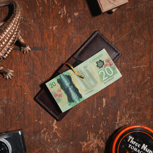 a full-grain dark brown leather money clip wallet open on a wooden table with a $20 Canadian bill surrounded by vintage props 