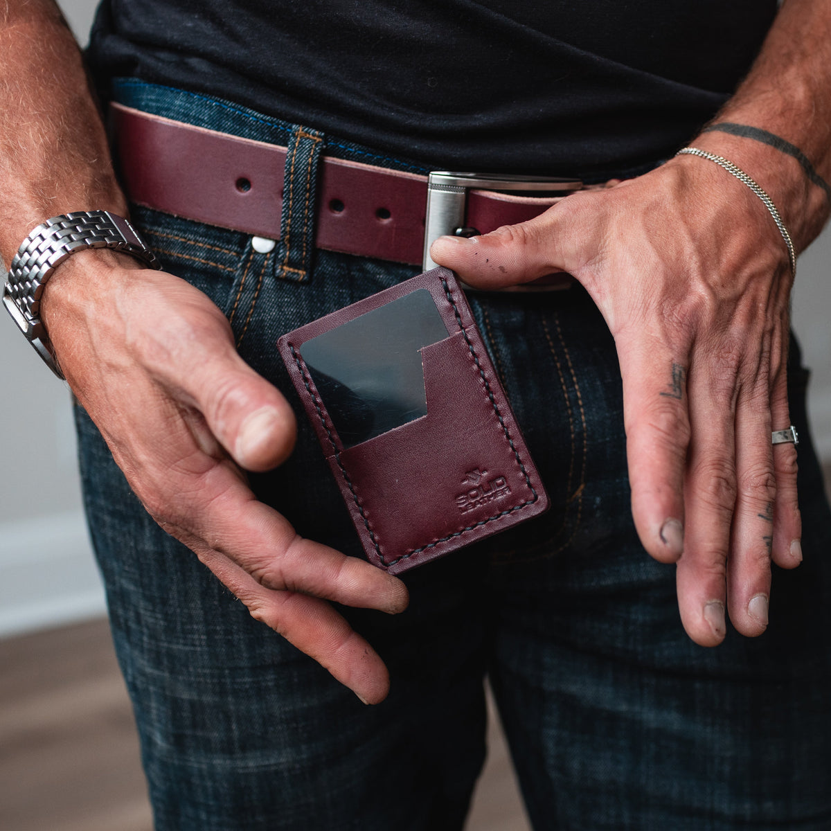 A pair of mans hands shows off a burgundy leather slim card holder wallet with matching burgundy leather belt on blue jeans and a black T