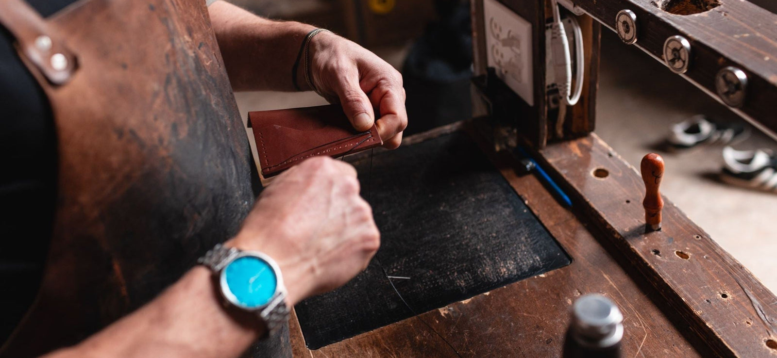 A close-up lifestyle shot of a leatherworker (Jesse Bateson) hand stitching a Sollid Leather wallet on his rustic wooden workbench. He is wearing a brown leather apron and a blue-faced watch.
