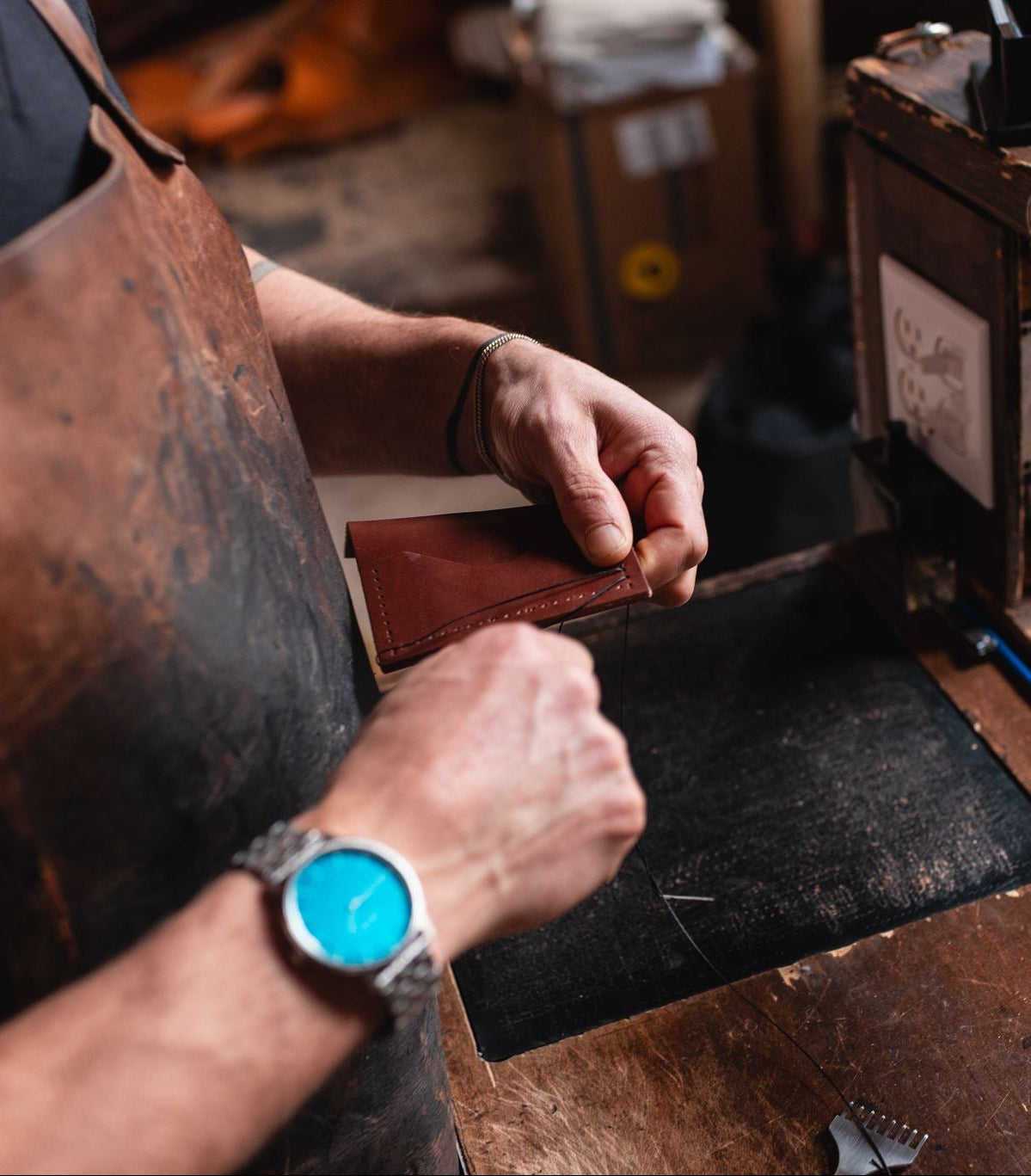 A close-up lifestyle shot of master leatherworker Jesse Bateson of Solid Leather hand stitching a full-grian leather wallet. He is wearing a brown leather apron and a blue-faced watch with rugged worker hands.