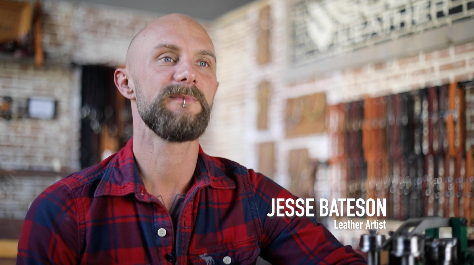 Good looking man in a plaid shirt with 'Jesse Bateson' text overlay in a leather workshop setting