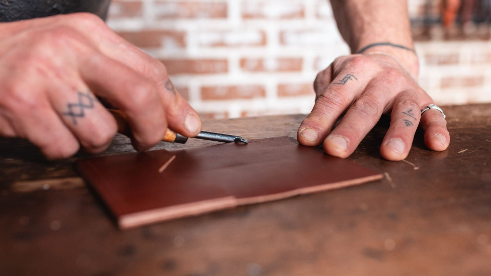 A close-up of Jesse Bateson's tattooed hands crafting a leather wallet on a wooden workbench at his Solid Leather studio in Peterborough Ontario Canada, with a blurred brick wall in the background.