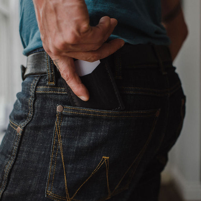 Close-up of a person wearing dark blue denim jeans with a black leather cardholder wallet partially pulled from the back pocket, set in a softly blurred indoor environment.