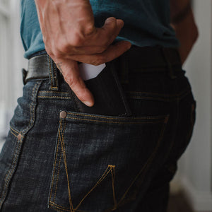 Close-up of a person wearing dark blue denim jeans with a black leather cardholder wallet partially pulled from the back pocket, set in a softly blurred indoor environment.