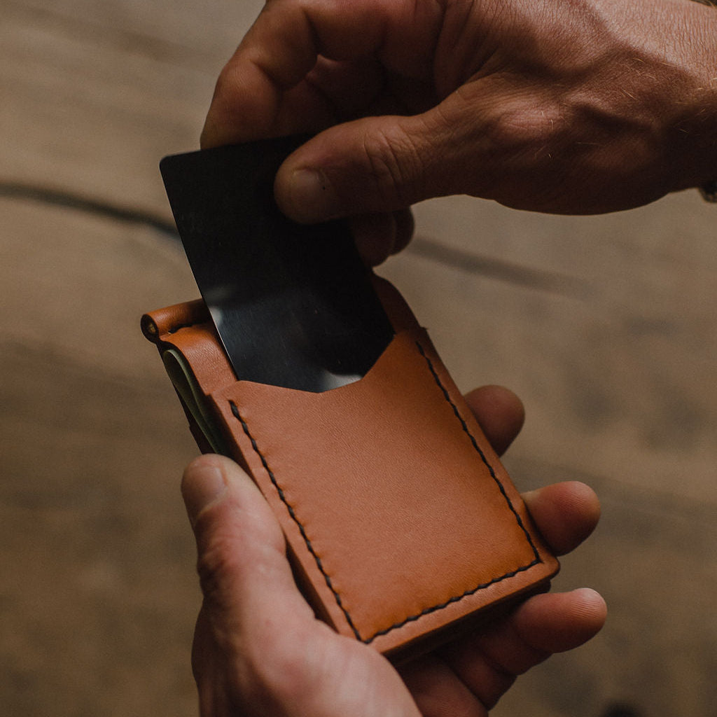 A close-up of a person's hands holding a slim, tan leather card holder wallet and inserting a black card into its front slip pocket, set against a blurred wooden background.