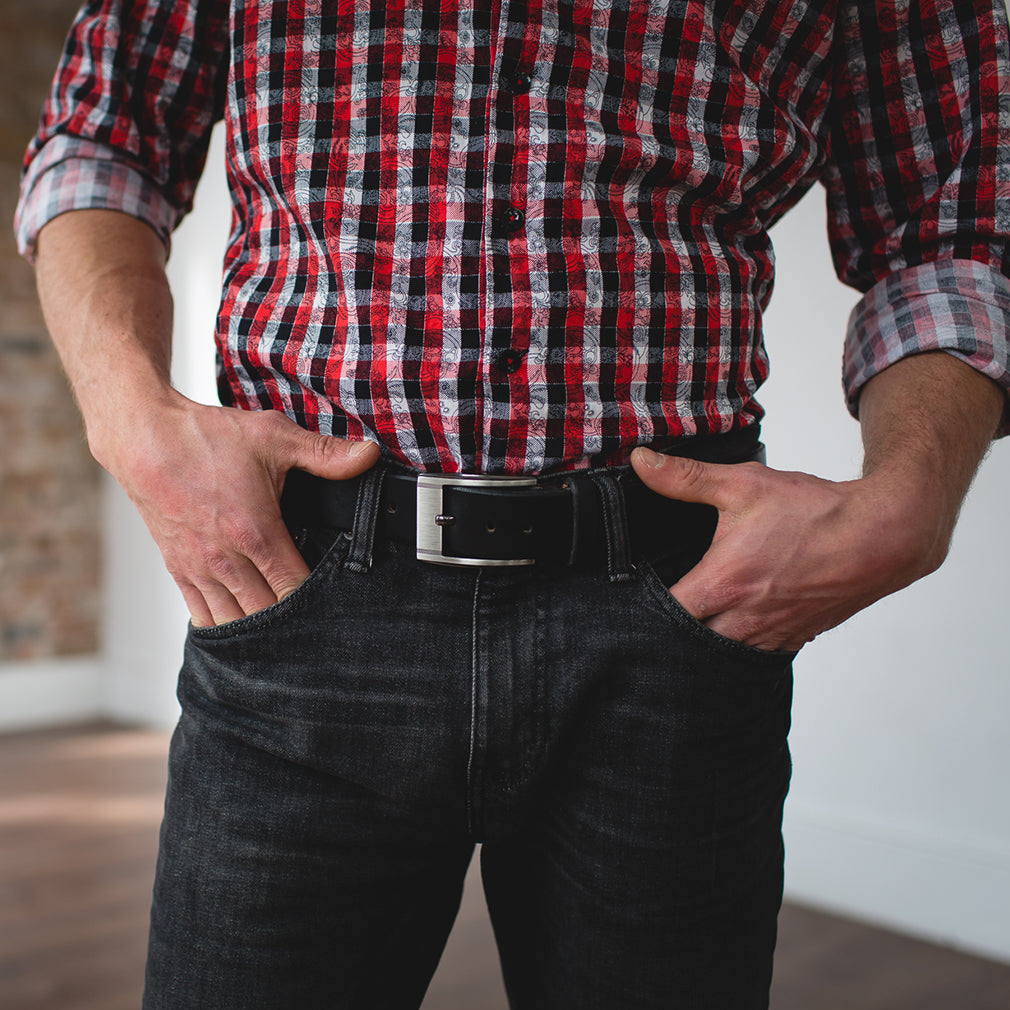 A man wearing a full-grain leather belt from solid leather in casual black with plaid shirt, black jeans and hands in pocket 
