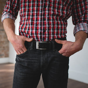 A man wearing a full-grain leather belt from solid leather in casual black with plaid shirt, black jeans and hands in pocket 
