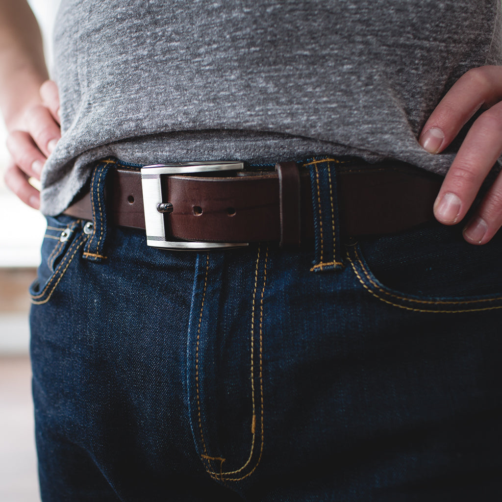 A full grain leather belt in dark brown with stainless steel buckle on blue jeans with grey t-shirt tucked in 
