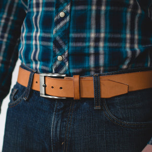 a tan full grain leather belt with stainless steel buckle and leather keeper worn on blue jeans with a blue plaid button up shirt 