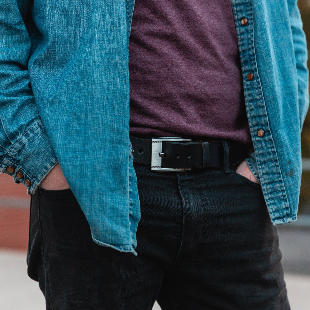 A person wearing a teal blue denim button-up shirt over a dark purple t-shirt, paired with black jeans and a black full grain leather belt with a silver buckle, standing outdoors.