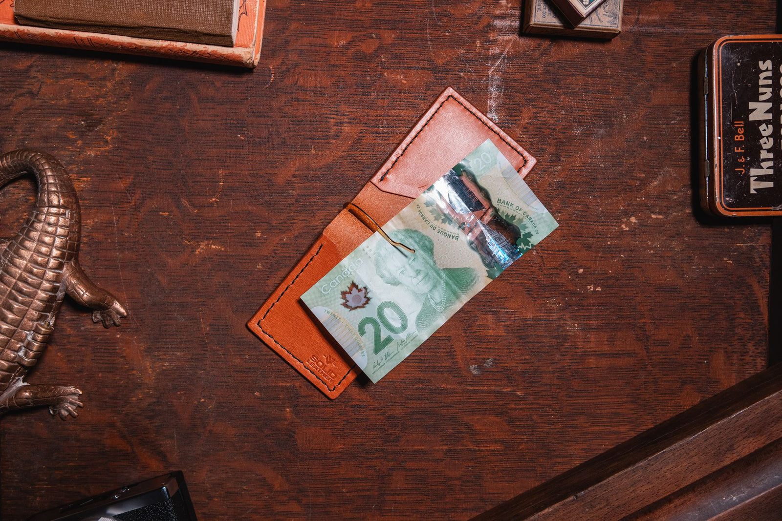 A tan brown leather cardholder wallet holding a Canadian 20-dollar banknote, placed on a dark wooden desk with vintage props.