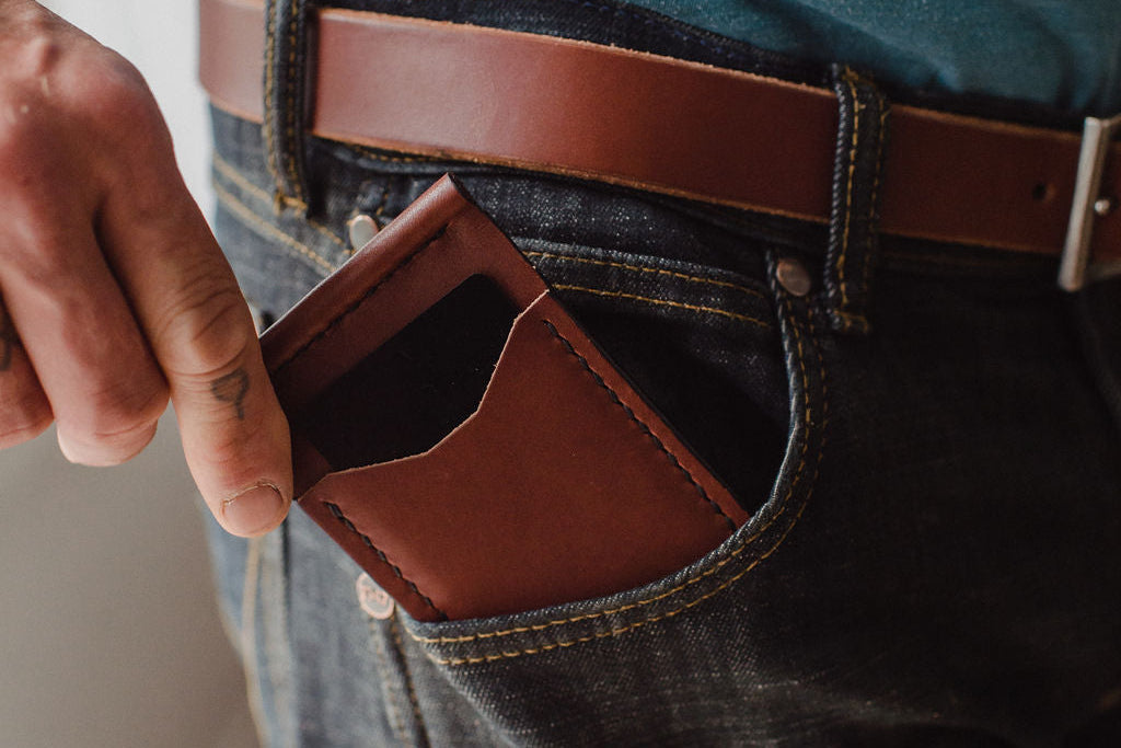 A close-up of a person wearing dark blue jeans and a cognac brown leather belt, with a matching brown leather card holder or wallet tucked into the front pocket.