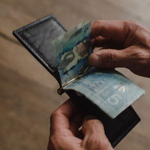 A close-up of a person's hands holding an open black leather wallet with Canadian five-dollar banknotes inside, set against a blurred wooden floor background.