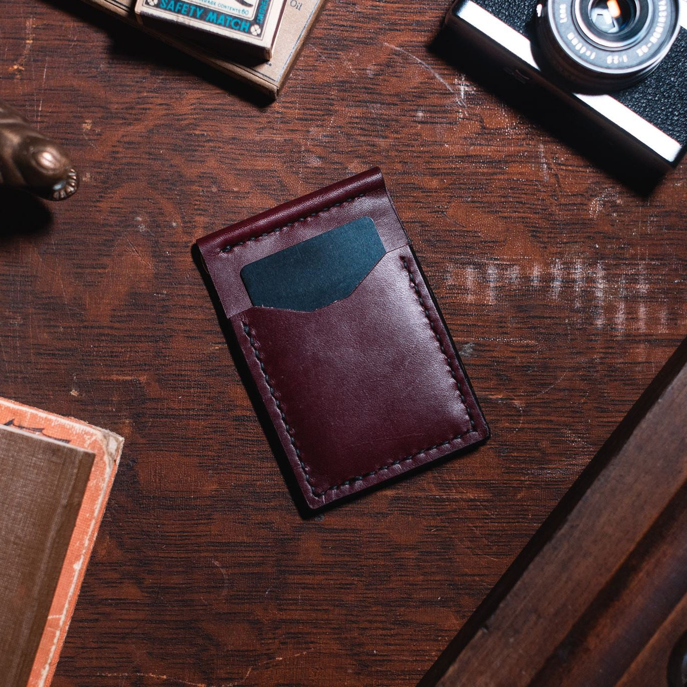 A rich burgundy leather card holder wallet with 4 card slots, shown on a dark wooden desk surrounded by vintage props including a camera, a brass alligator figurine, and a book.