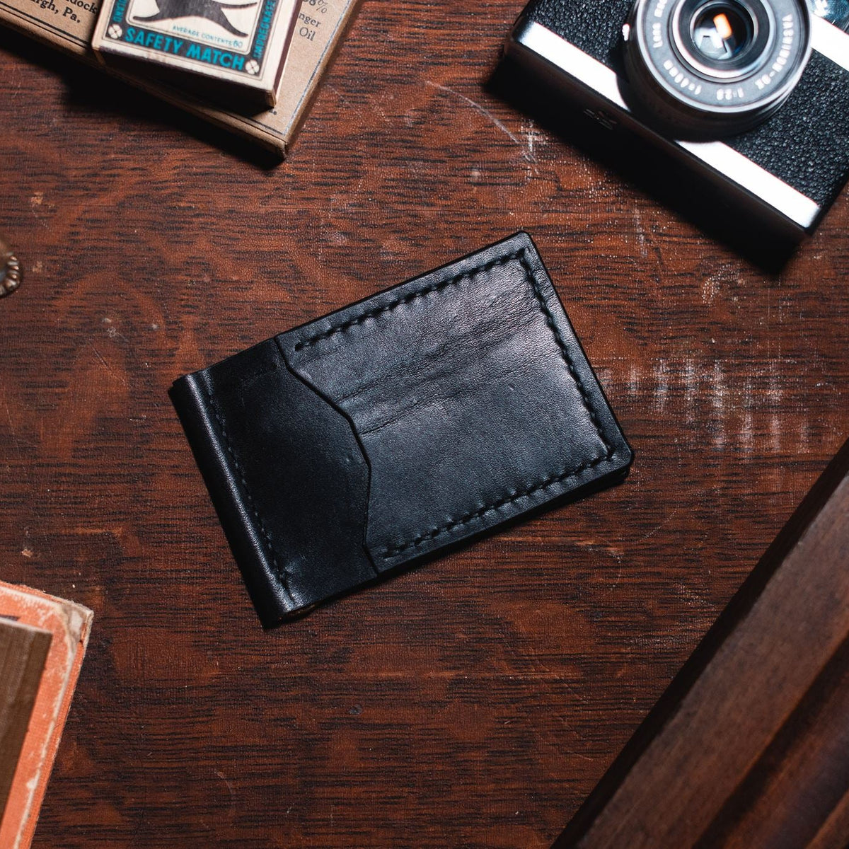 A black leather card holder wallet with visible stitching, placed on a dark wooden tabletop surrounded by vintage props including a camera, books, and a brass object.