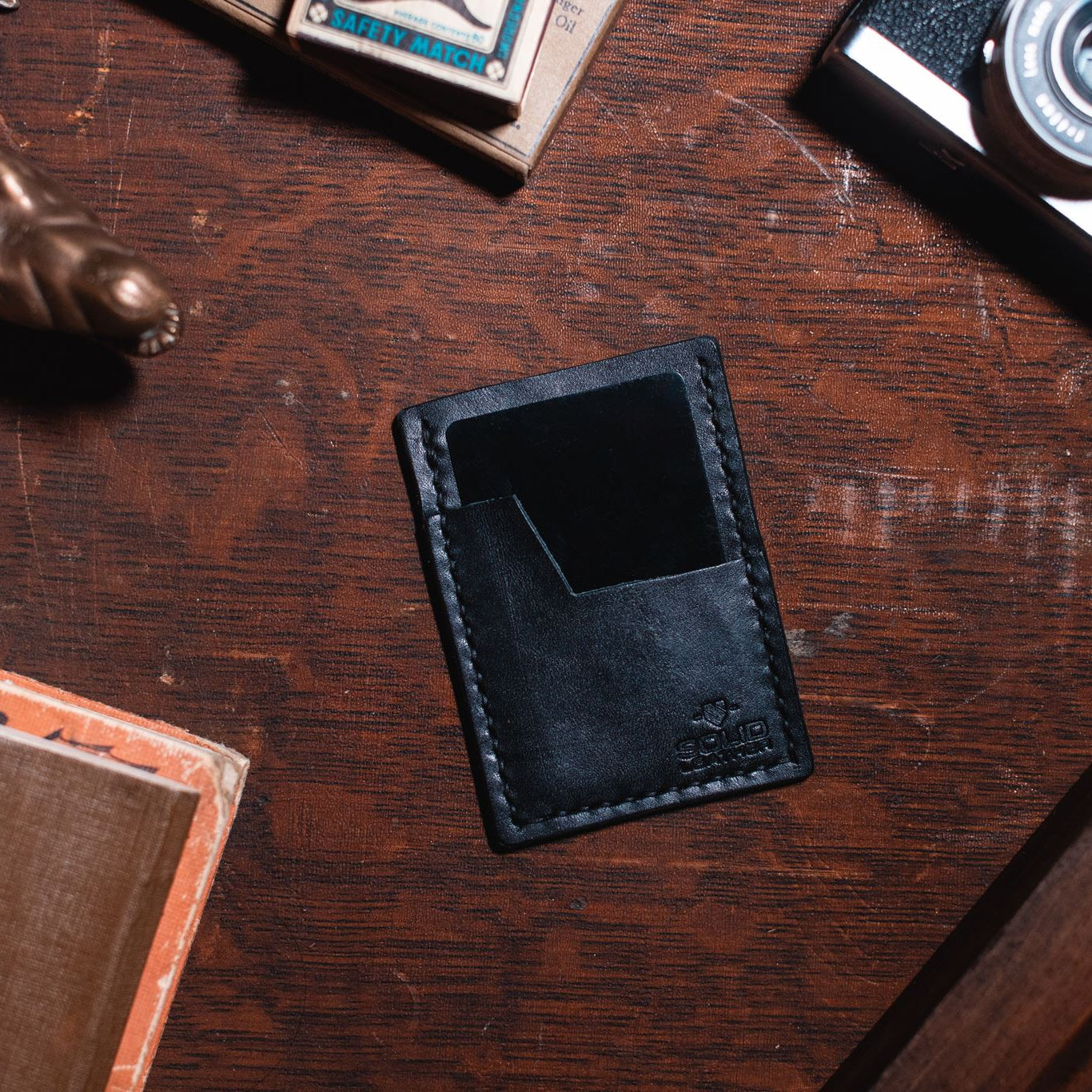 A black leather card holder wallet with a slanted card pocket, shown from above on a dark wooden desk surrounded by vintage props including a camera, a small figurine, and a book.