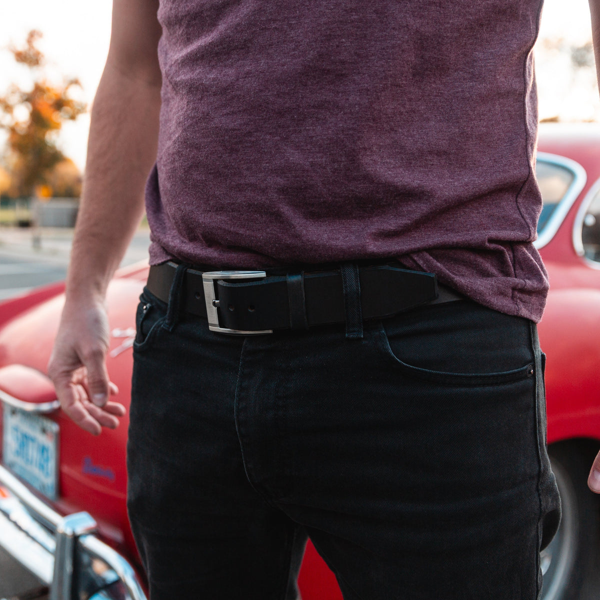 Casual black full-grain leather belt on dark jeans with a blurred red car in the background   