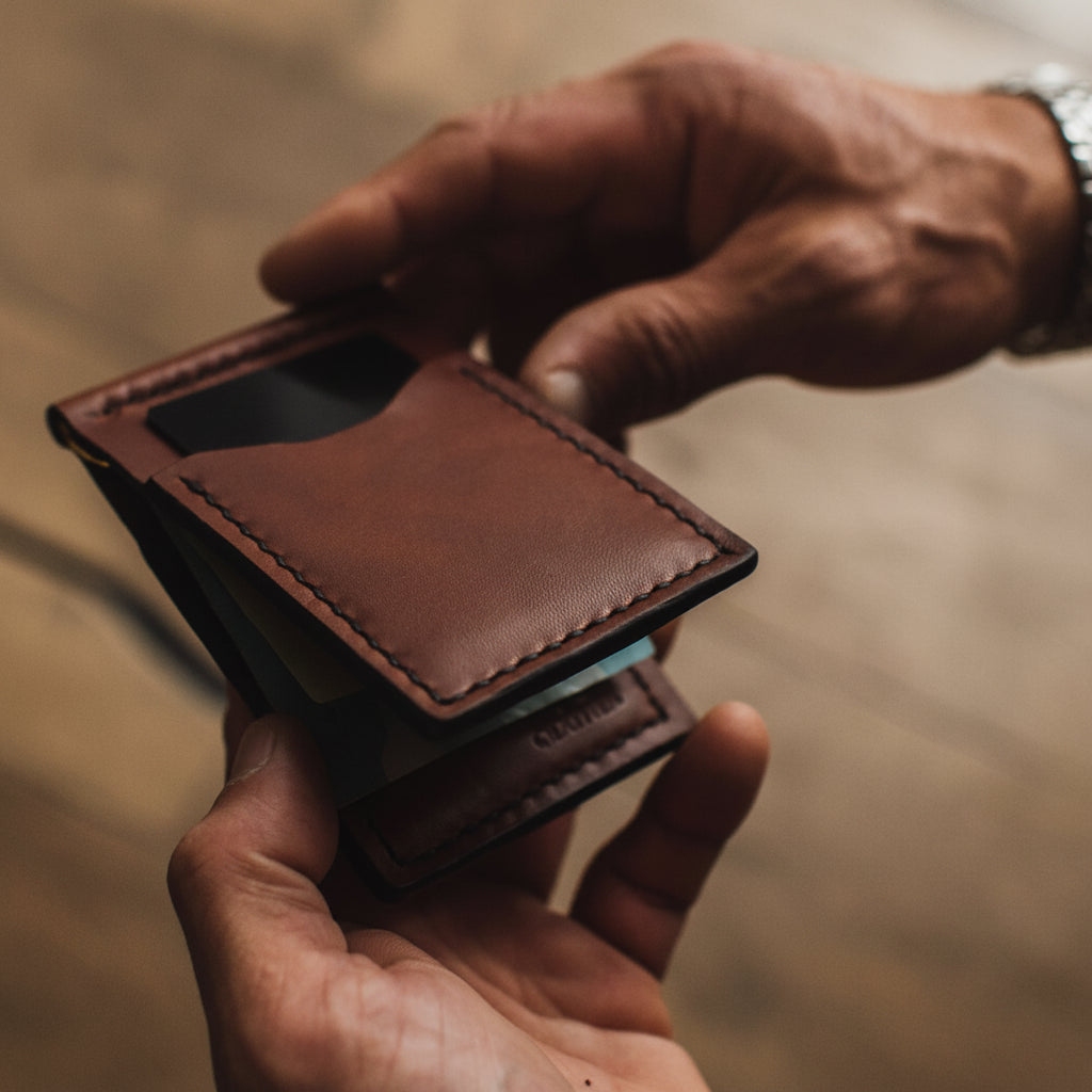 A close-up of a person's hands holding an open cognac brown leather card holder wallet, showing its interior compartments and card slots, with a blurred warm-toned background.