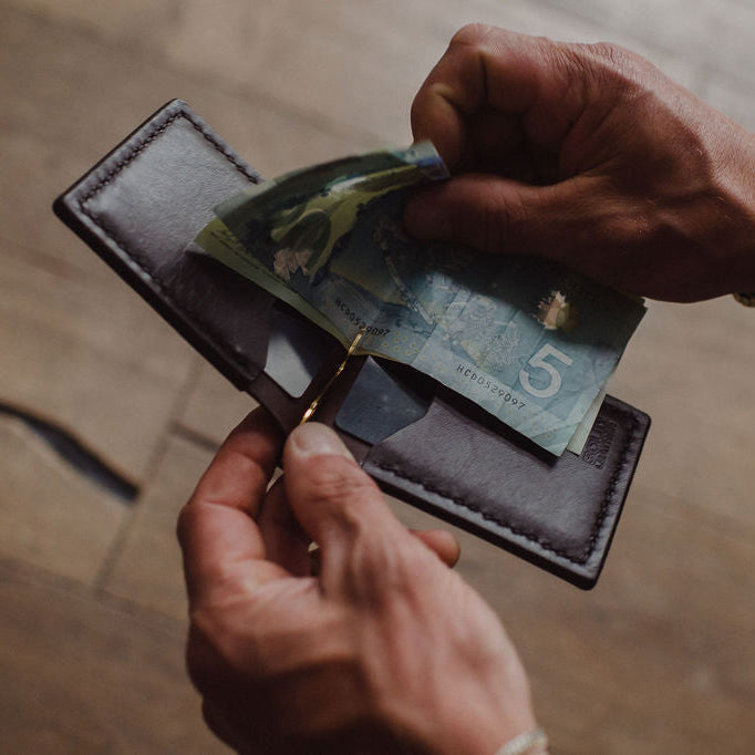 A close-up of a person's hands holding an open dark brown solid leather wallet, inserting a Canadian five dollar bill , with a gold-tone card or ID holder in the center pocket, set against a blurred wooden floor background.