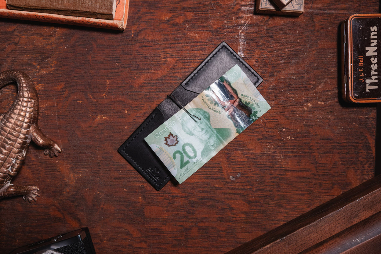 A Black leather cardholder wallet holding a Canadian 20-dollar banknote, placed on a dark wooden desk with vintage props.