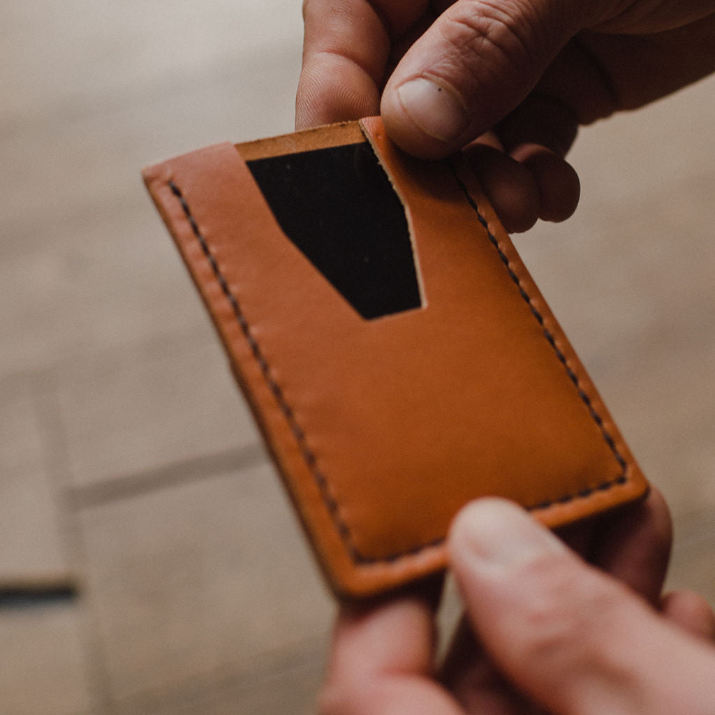 A close-up of a person's hands holding a minimalist tan leather card holder wallet, set against a blurred wooden background.