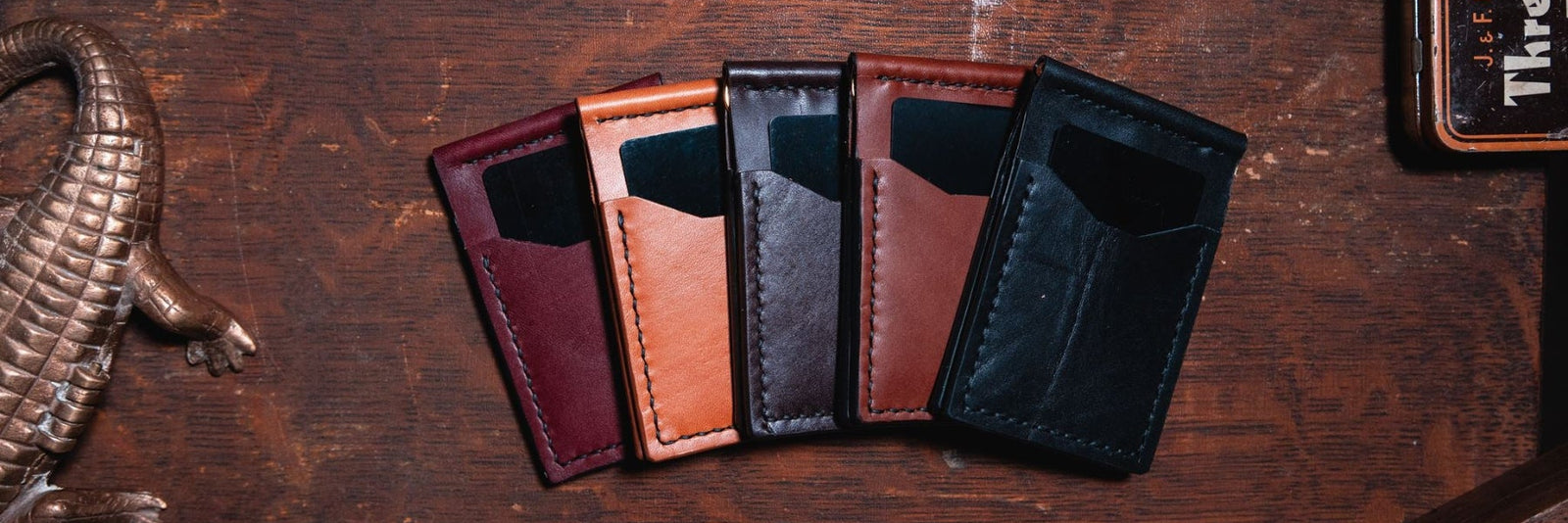 Five slim leather card holders money clip wallets in different colours (burgundy, tan, dark brown, cognac brown, and black) arranged in a fan shape on a dark wood tabletop.