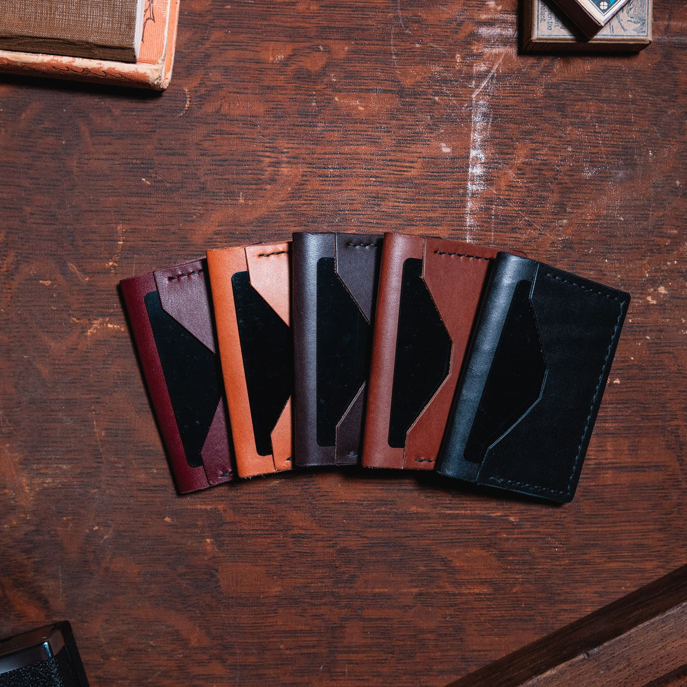 Solid Leather everyday bifold wallet in five leather colours. Black, cognac brown, dark brown, tan and burgundy, on a dark wooden desk. 