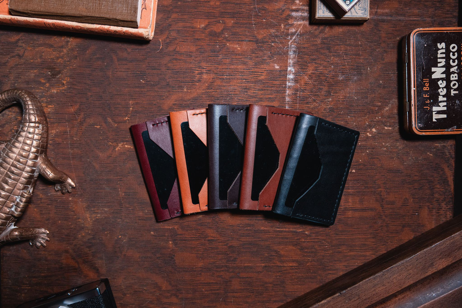 five Solid Leather everyday bifold wallets in black, cognac brown, dark brown, tan and burgundy on a dark wooden desk. Handmade in Peterborough Ontario by Jesse Bateson of Solid Leather 