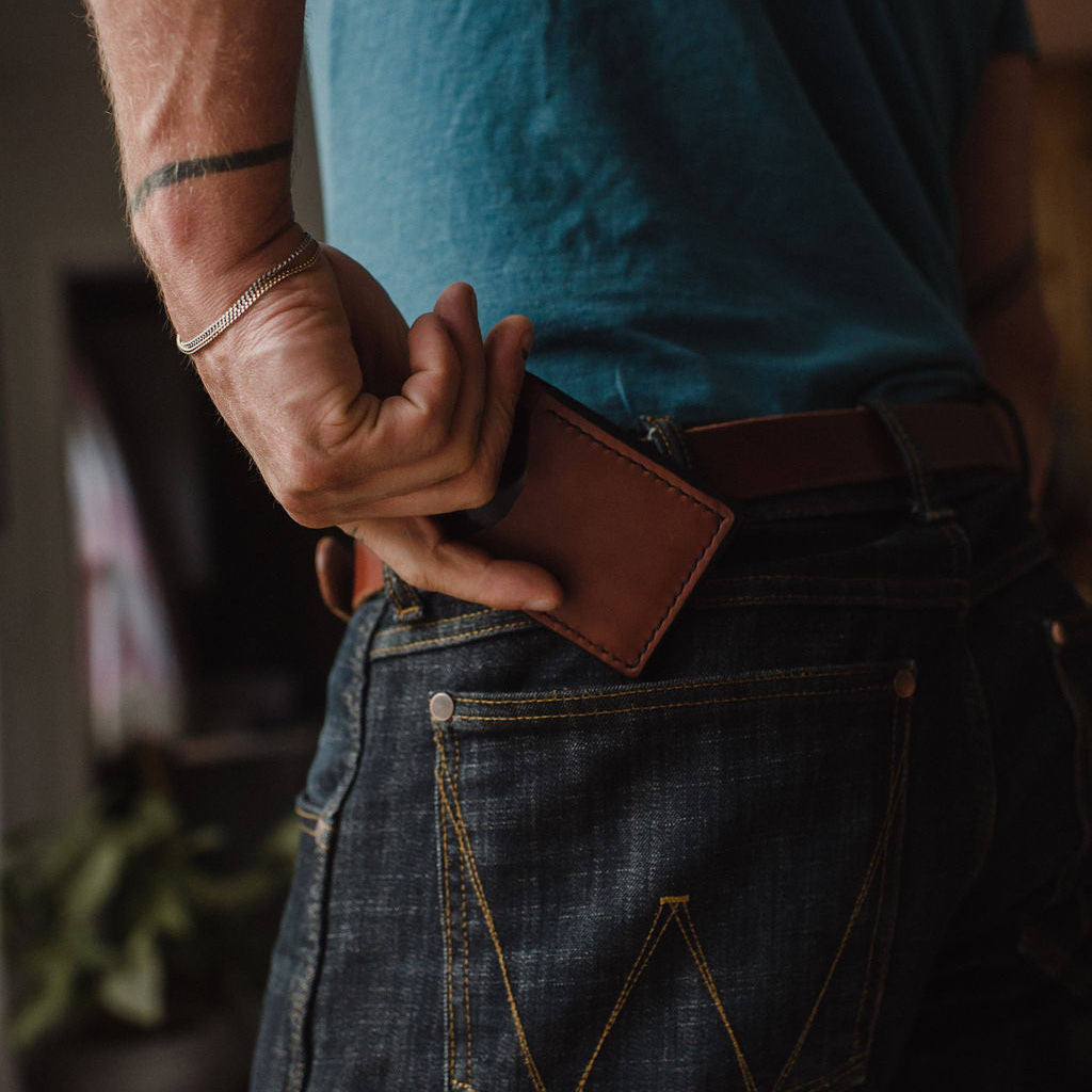 a Solid Leather cognac brown bifold cardholder wallet being inserted into a pair of dark blue jeans being held up by a matching Solid Leather belt 