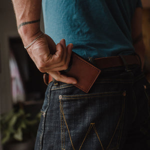 a Solid Leather cognac brown bifold cardholder wallet being inserted into a pair of dark blue jeans being held up by a matching Solid Leather belt 