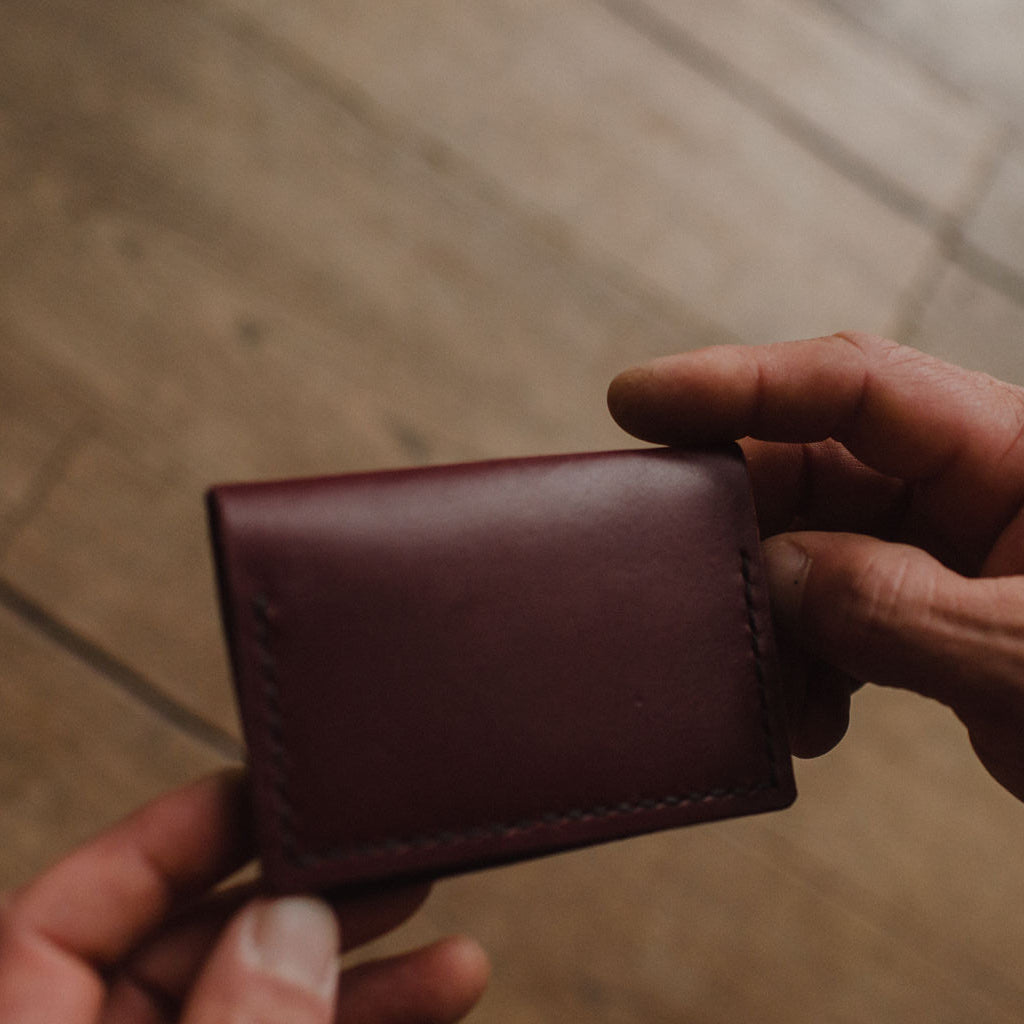 A close-up of a person's hands holding a burgundy leather bifold cardholder wallet against a blurred wooden floor background.