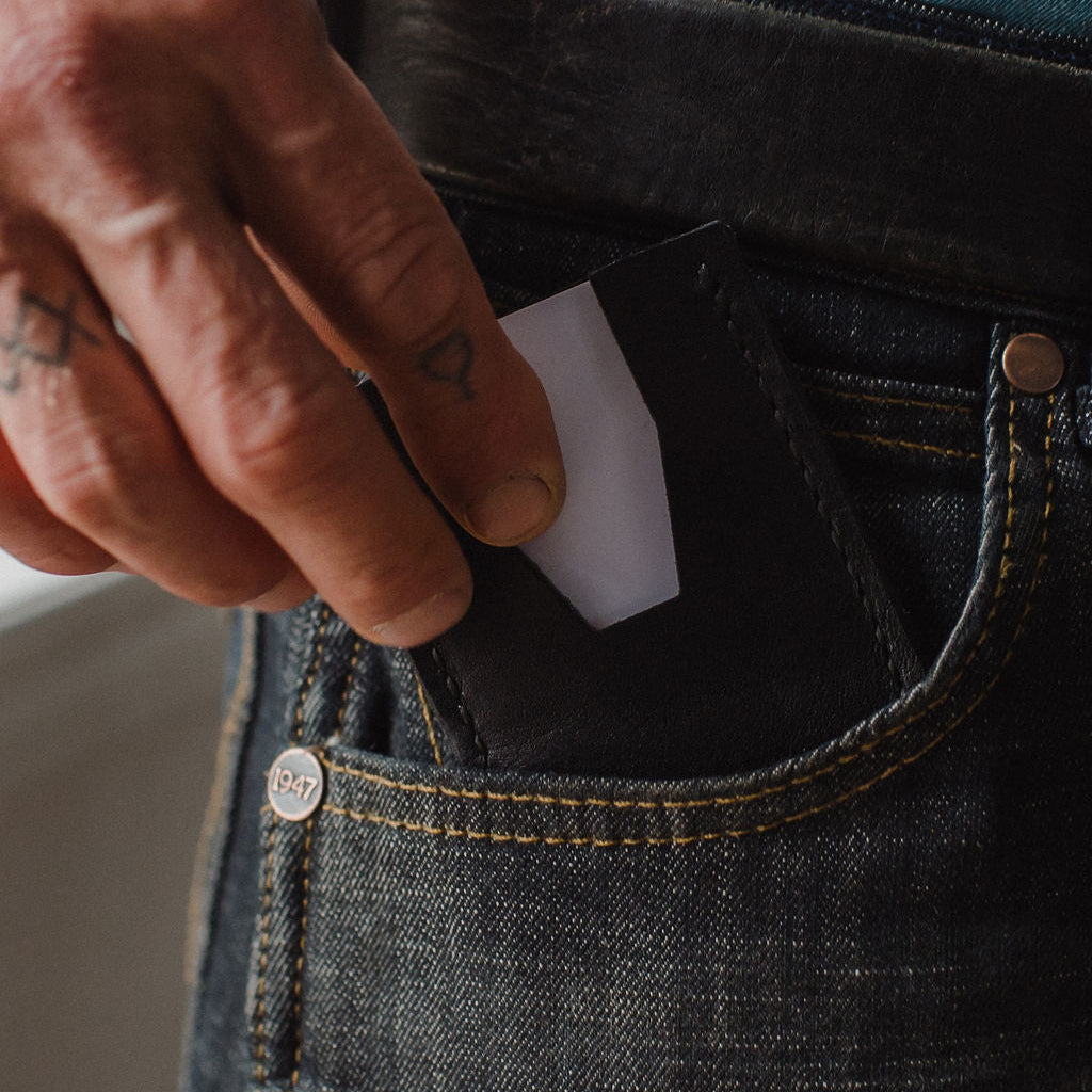 A close-up of a person's hand inserting a black leather cardholder wallet into the pocket of dark wash denim jeans.