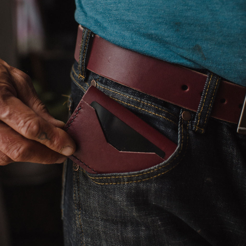A close-up of a person wearing a burgundy leather card holder in their jeans pocket, paired with a blue t-shirt and a matching burgundy solid leather belt.