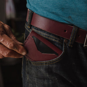 A close-up of a person wearing a burgundy leather card holder in their jeans pocket, paired with a blue t-shirt and a matching burgundy solid leather belt.
