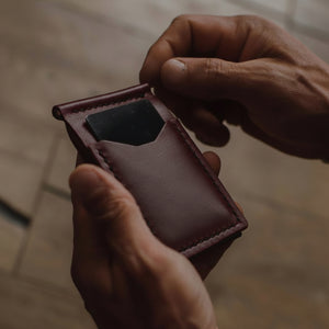 A close-up of a person's hands holding a  burgundy/oxbloodleather card holder wallet against a blurred wooden background. Handmade by Jesse Bateson at his Solid Leather workshop in Peterborough Onatrio