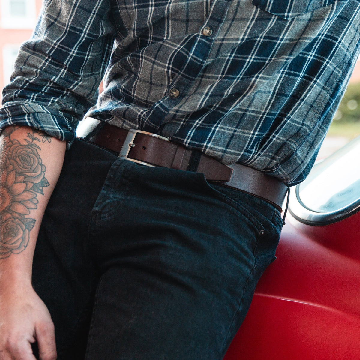 a man leans against a red car wearing a full grain Solid Leather belt in brown with dark jeans and a plaid shirt 