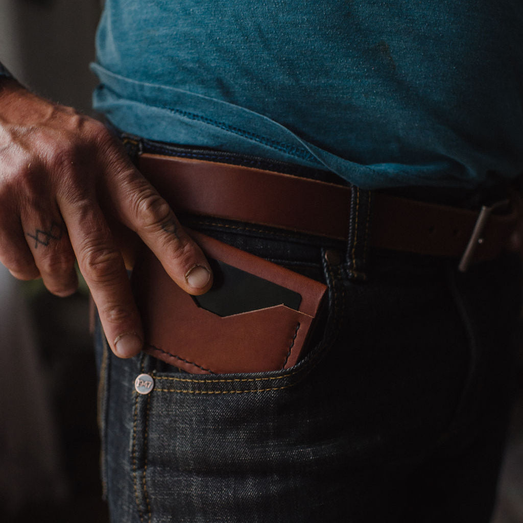 a slim cognac brown leather bifold cardholder wallet being inserted into a pair of dar blue jeans eing held up by a matching Solid Leather belt