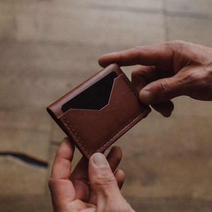a pair of mans hands holding a cognac brown leather bifold cardholder wallet showing the backside card sleeve on a blurred background