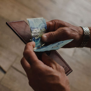 A close-up of a person's hands holding an open cognac brown leather wallet with Canadian banknotes inside, against a blurred wooden floor background.