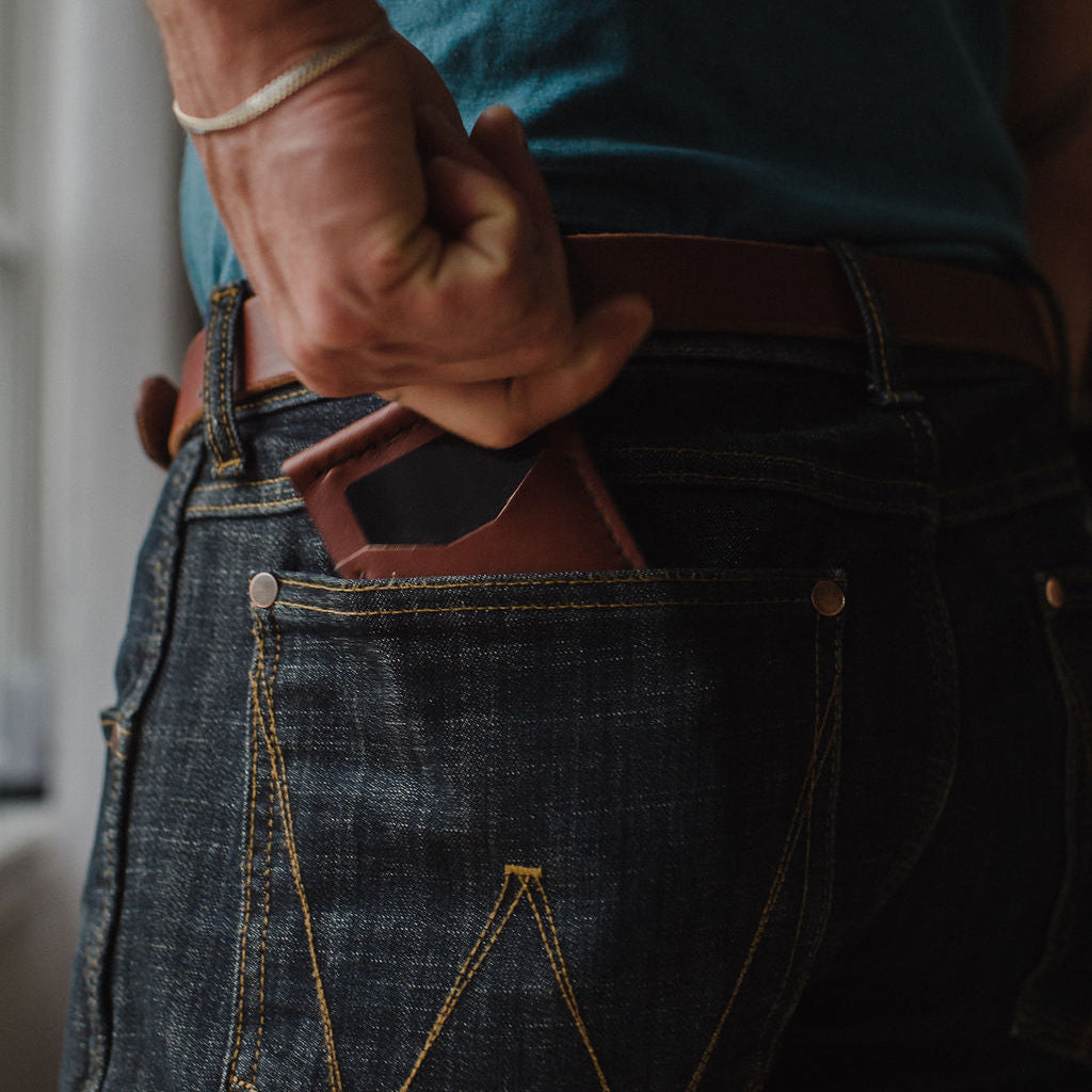 Close-up of a person wearing dark blue denim jeans with a Cognac brown leather belt and a matching brown leather money clip wallet in the back pocket.