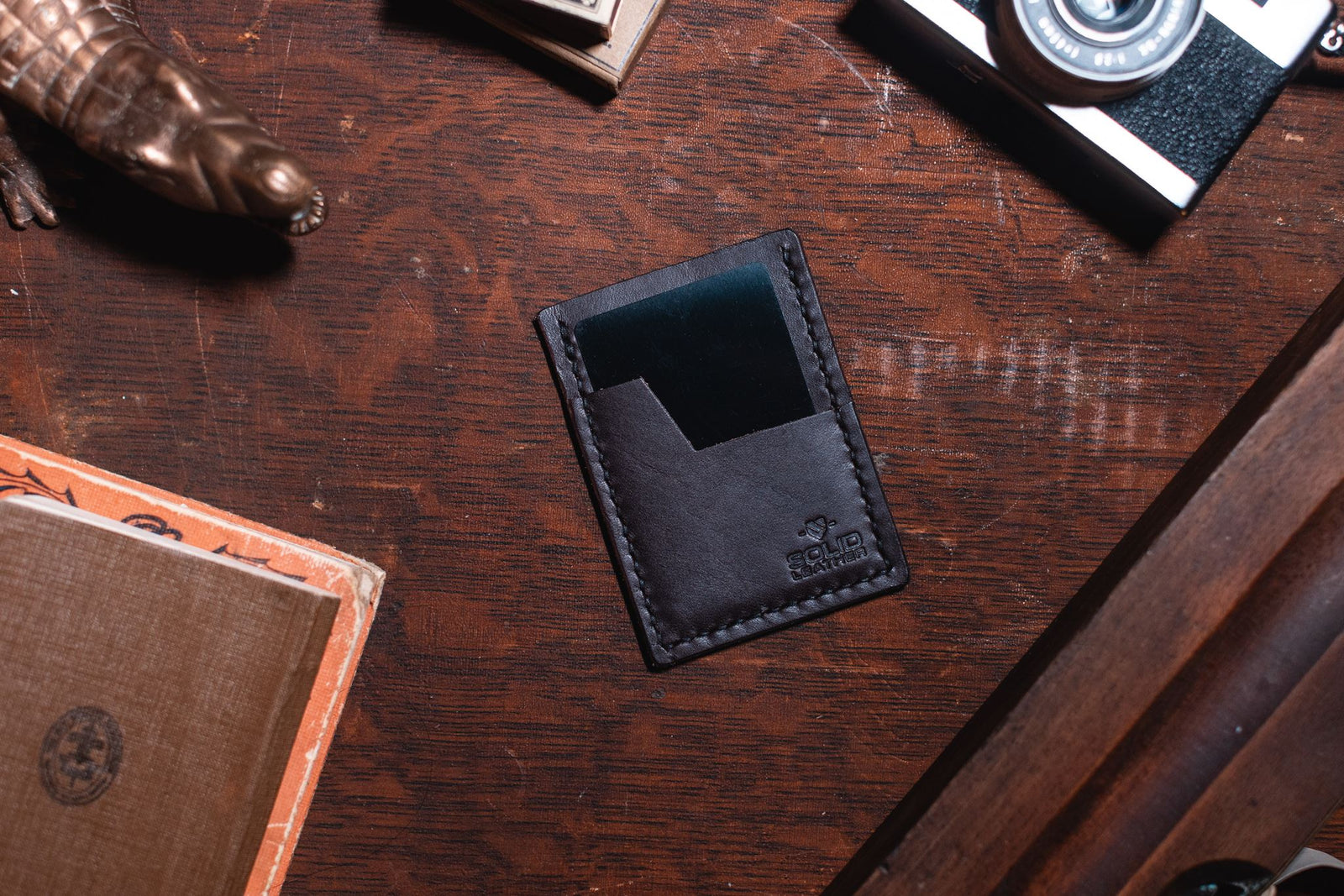a dark brown minimalist wallet on a dark wooden desk surrounded by vintage props. handmade in Peterborough Ontario by Jesse bateson of Solid Leather 