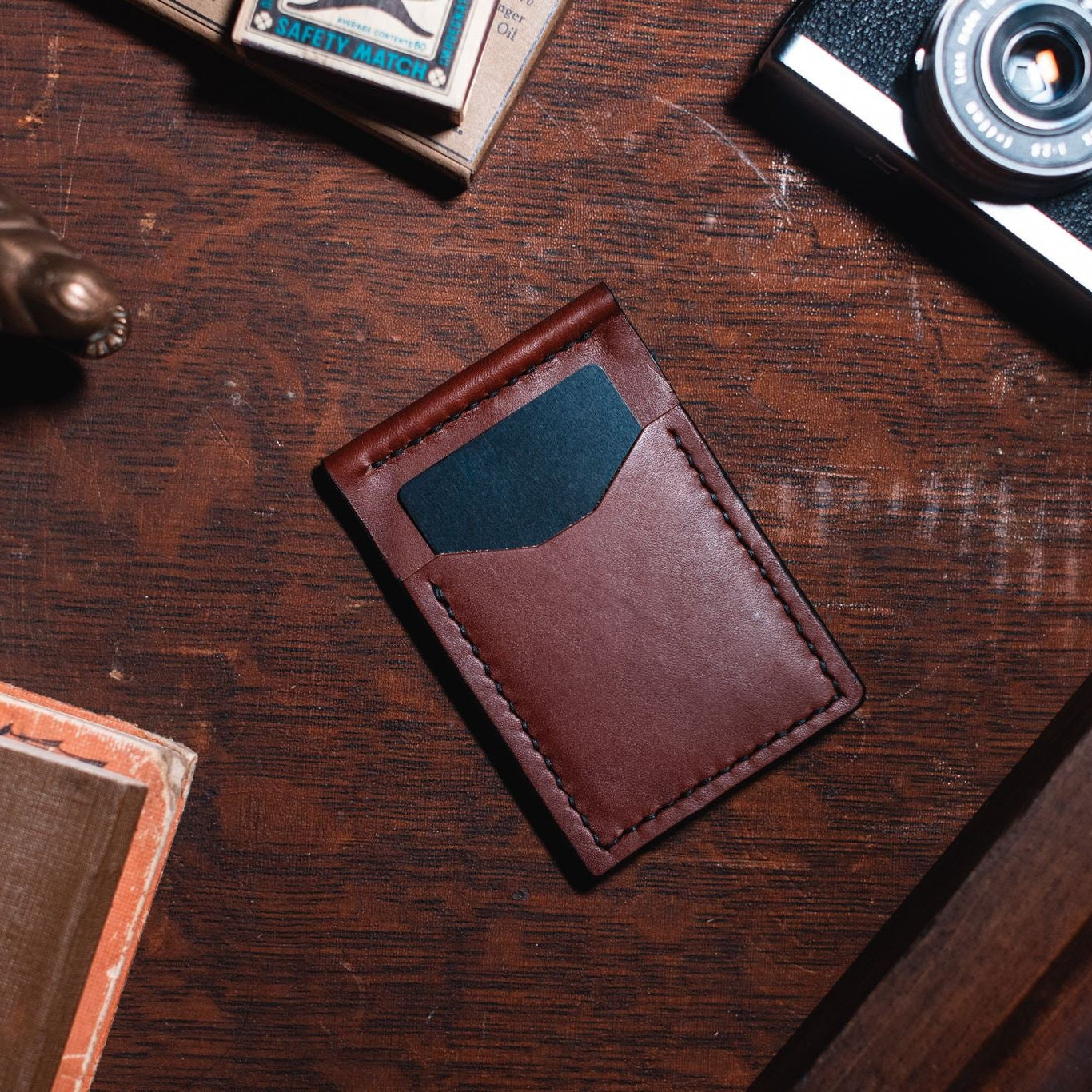 A cognac brown leather card holder wallet with 4 card slots, placed on a dark wooden desk surrounded by vintage props including a camera, a small figurine, and papers. Handmade in Peterborough Onatrio Canada by jesse bateson of Solid Leather 